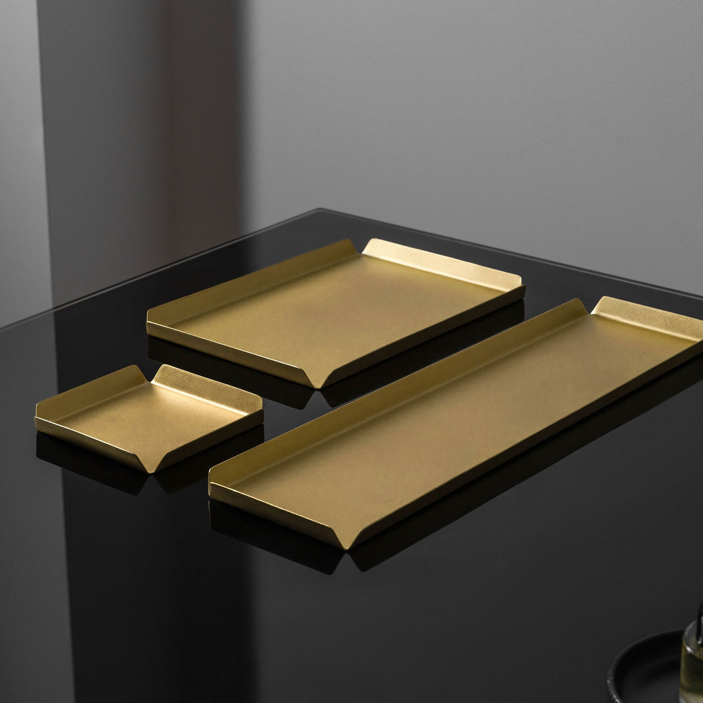 A small, square shaped brass tray, behind that is a medium shaped brass tray and to the right of both of those trays is a long, rectangle shaped brass tray. All the trays have raised edges. The trays are photographed upon a black reflective surface. The brass trays are made by Empire Copper.