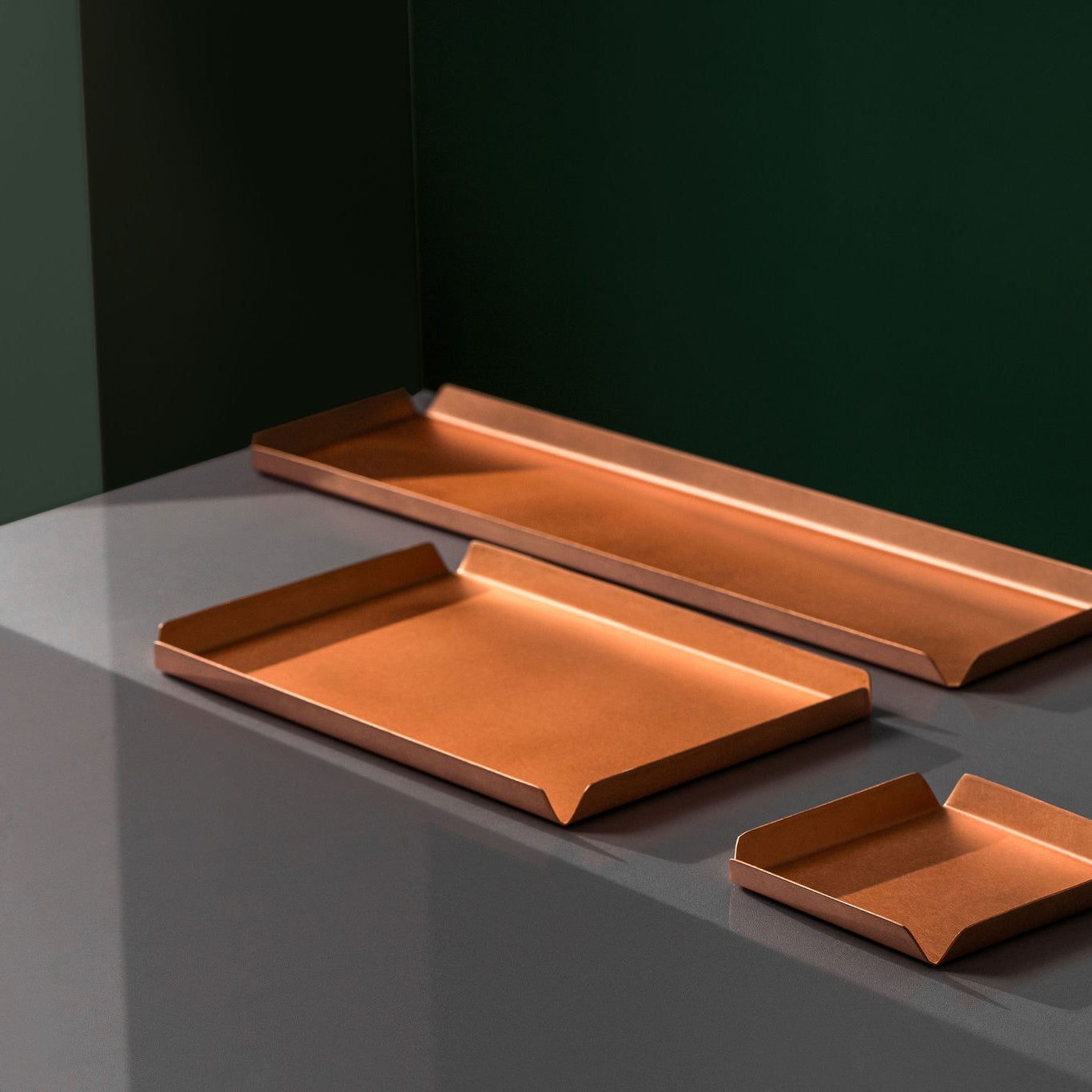 A 100mm x 100mm square copper tray, a 235mm W x 150mm D copper tray and a 400mm W x 100mm D copper tray all sitting upon a white surface. The background of the image is emerald green. The copper trays are made by Empire Copper.