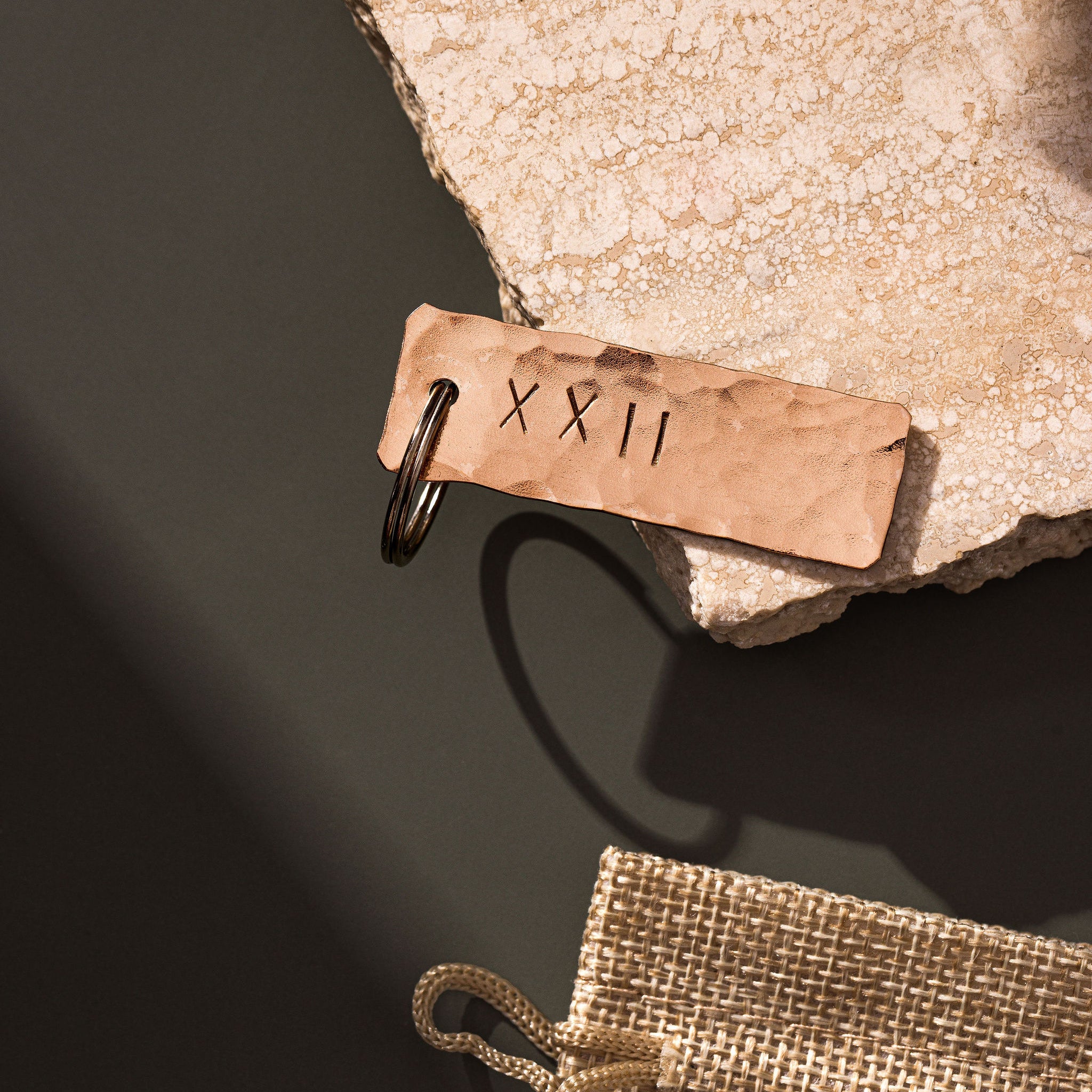 A copper keyring that has been planished and engraved with the roman numeral symbols XXII which translates to the number 22 sitting atop a rugged piece of travertine. The travertine slab is laid upon a moss green background. The copper keyring is made by Empire Copper.