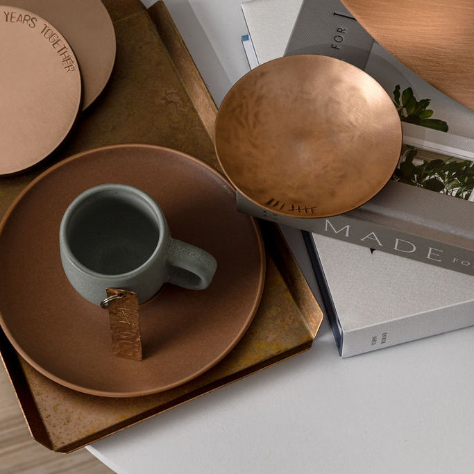 A small, round, bronze trinket dish with the tally number eight engraved on the left handside just inside the rim. To the left of the dish there is a trinket tray with a white teacup handle barely in the frame. The trinket dish is sat upon a stack of grey and white home and lifestyle books. The trinket dish is made by Empire Copper.