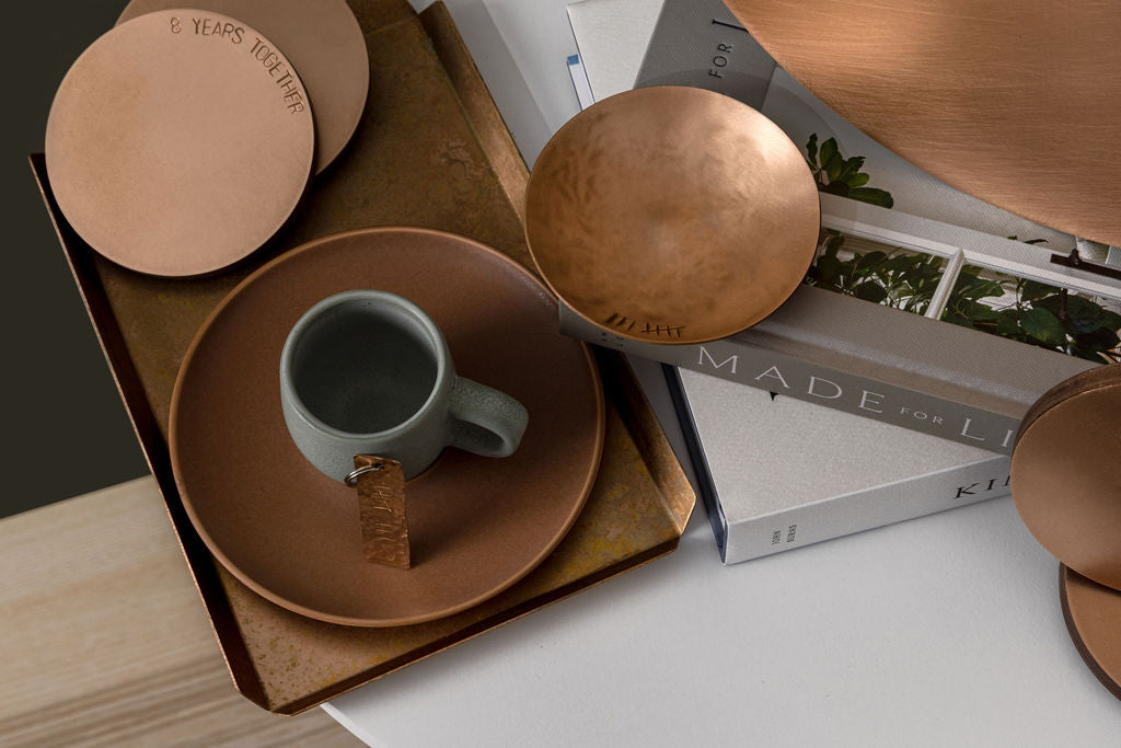 A small, round, bronze trinket dish with the tally number eight engraved on the left handside just inside the rim. To the left of the dish there is a trinket tray with a white teacup handle barely in the frame. The trinket dish is sat upon a stack of grey and white home and lifestyle books. The trinket dish is made by Empire Copper.