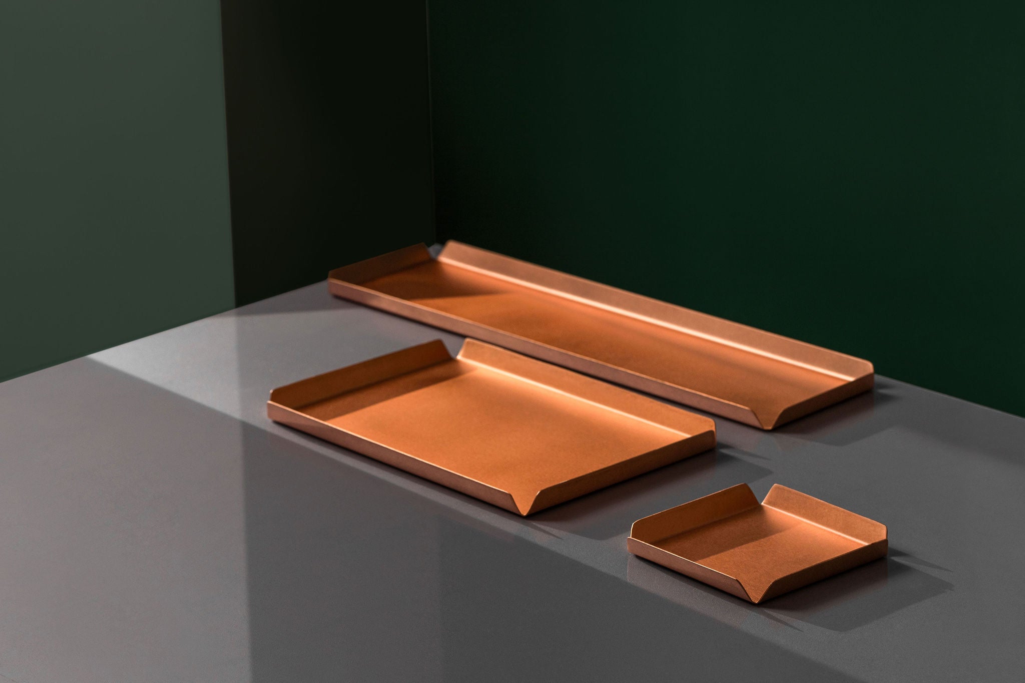 A 100mm x 100mm square copper tray, a 235mm W x 150mm D copper tray and a 400mm W x 100mm D copper tray all sitting upon a white surface.  The background of the image is emerald green. The copper trays are made by Empire Copper.